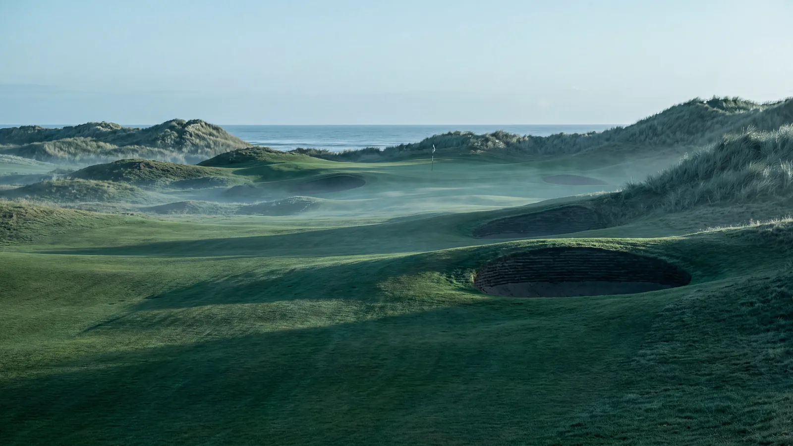 Dawn mist lying in the hollows of a links golf hole, a deep revetted pot bunker in the foreground and the sea beyond the green